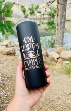 a person holding up a black one happy camper tumbler in front of a river