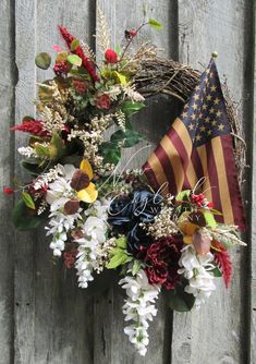 a wreath with an american flag hanging on a wooden fence next to flowers and foliage