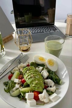 a white plate topped with salad next to a glass of green juice and a laptop