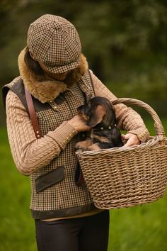 a woman holding a basket with a dog in it while wearing a hat and coat