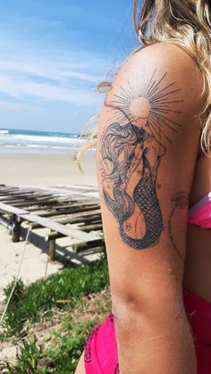 a woman with a mermaid tattoo on her arm and shoulder, standing in front of the ocean