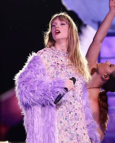 two women are performing on stage with one holding a microphone and the other wearing a purple feather coat