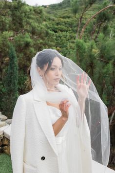 a woman wearing a white veil and holding her hand up in the air while standing next to a lush green hillside