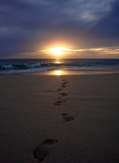 the sun is setting over the ocean with footprints in the sand