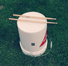 a white bucket with two wooden sticks sticking out of it sitting in the grass next to a pair of drumsticks