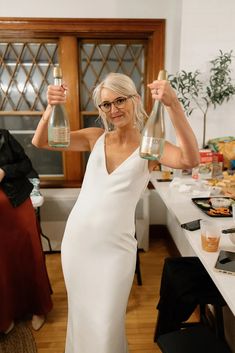 a woman in a white dress holding up two wine bottles and posing for the camera