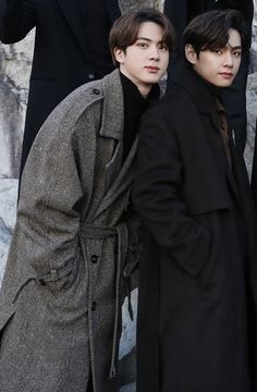 two young men standing next to each other in front of a stone wall wearing coats