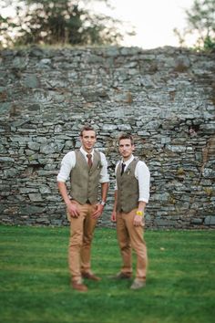 two men standing next to each other in front of a stone wall and green grass