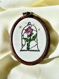 a cross stitched picture of a pink rose in a glass dome on white fabric