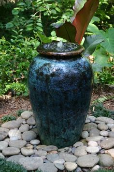 a large blue vase sitting on top of a pile of rocks next to a lush green bush