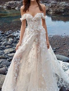 a woman in a wedding dress standing on rocks near the water with her hands behind her back