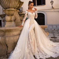 a woman standing in front of a fountain wearing a white wedding dress with long sleeves