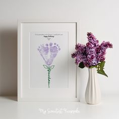 a white vase with purple flowers in it next to a framed print that says happy mother's day