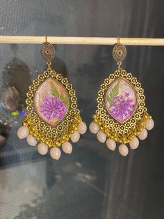 These handmade resin earrings feature a vibrant purple flower encased in a beige background, complemented by gold-earring and dangling beads. Perfect for adding a boho-chic touch to any outfit. Unique Flower-shaped Resin Earrings, Bohemian Pressed Flowers Drop Earrings, Pink Bohemian Resin Jewelry, Bohemian Pink Resin Jewelry, Dangle Earrings With Flower Charm In Resin, Bohemian Style Resin Flower Earrings As Gift, Purple Resin Jewelry With Matching Earrings, Bohemian Gold Resin Earrings, Resin Drop Earrings With Birth Flower Design