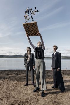 three people standing on the beach holding up a chess board with birds flying above them