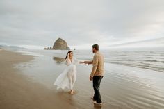 a man and woman holding hands on the beach near an ocean with a rock in the background