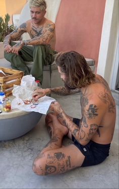 two men sitting on the ground with tattoos on their body and one is eating food