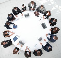 a group of people sitting around a white table with laptops and papers on it