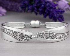 two silver bracelets with flowers on them and purple flowers in the backgroud