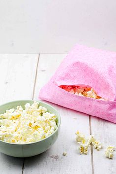 a bowl of popcorn next to a pink bag on a white wooden table with the lid open