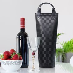 a wine bottle, glass and strawberries on a white table with a black checkered tote bag