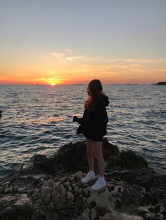 a woman standing on top of a rocky cliff next to the ocean at sunset or dawn