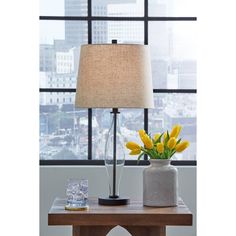 a table with a lamp, vase and flowers on it in front of a window