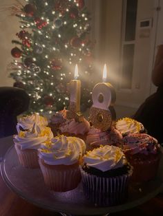 cupcakes with white frosting and gold numbers are on a cake plate in front of a lit christmas tree