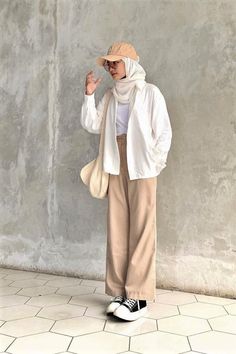 a woman standing in front of a concrete wall wearing a white jacket and beige pants