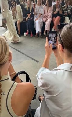 two women taking pictures with their cell phones in front of an audience at a fashion show