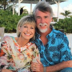 a man and woman sitting next to each other on top of a sandy beach with palm trees in the background