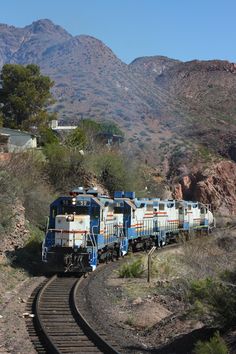 a blue and white train traveling down tracks next to mountain side covered in greenery