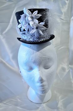 This hat is excellent for making a statement, its neautral colors allow you to build it into a focal point with any outfit. This satin miniature top hat can be worn with a headband or affixed to the wearers head via small loops inside the hat. Delicate white lace makes up the band, creating a stark contrast with the satin hat and forming a generous bow and waterfall tail in the back. The front of the band is decorated with a line of large rhinestones. Smaller rhinestones scatter thier glittery light around the rest of the hat. The front of the hat features small white velvet poinsettia flowers, with small gear and cog centers. The side features white feathers with a black and white cameo. Whimsical Costume Hat With Adjustable Round Crown, Whimsical Adjustable Costume Hat With Round Crown, Brimmed Party Costume Hats And Headpieces, Whimsical Mini Hats For Parties, Handmade Fitted High Crown Mini Hats, Handmade Fitted Mini Hat With High Crown, Party Brimmed Top Hat, Adjustable Round Crown Mini Hats For Kentucky Derby, Whimsical Fitted Top Hat With Short Brim