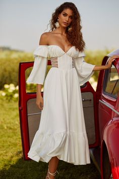 a woman standing next to a red car wearing a white dress with ruffles