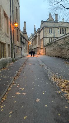 two people walking down the street in an old town at dusk with autumn leaves on the ground