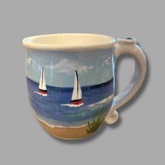 a coffee cup with two sailboats painted on the side and in front of an ocean scene