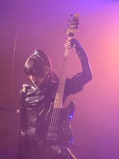 a woman in black leather outfit holding up a guitar on stage with purple lighting behind her