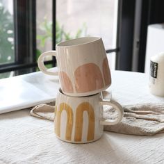 two mugs sitting on top of each other on a table next to a window