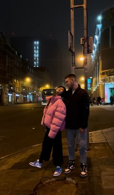 a man and woman standing next to each other on a street corner at night with buildings in the background