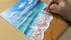 a person is drawing on a piece of paper with water and clouds in the background