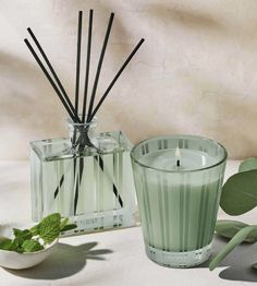 a candle and some green leaves on a white table with a glass container filled with reeds