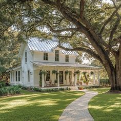 a white house sitting under a large tree on top of a lush green field next to a sidewalk