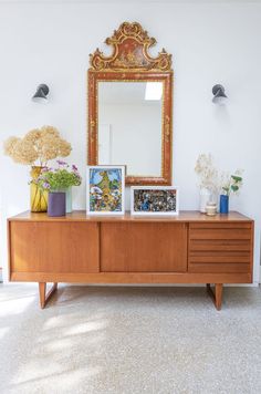 a wooden dresser sitting next to a mirror on top of a white wall with flowers in vases