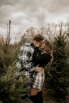 a man and woman kissing in front of christmas trees at an outdoor tree farm on a cloudy day