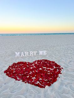 a heart made out of flowers sitting in the sand at the beach with words marry me written on it