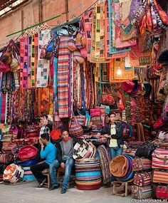 people are sitting in front of an outdoor market with colorful fabrics and rugs on display