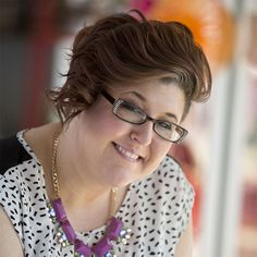 a woman wearing glasses and a purple necklace smiles at the camera while looking down on her cell phone