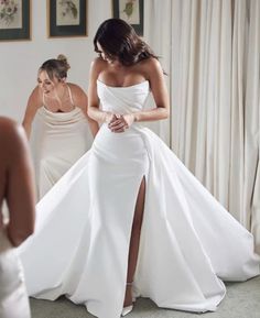 a woman in a white wedding dress is looking at herself in the mirror while another woman looks on