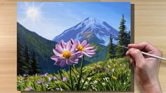 a painting of a mountain with flowers in the foreground and a person's hand holding a paintbrush