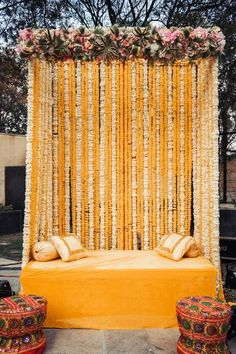 a bed with pillows and flowers on it in front of a yellow curtain that is hanging from the ceiling